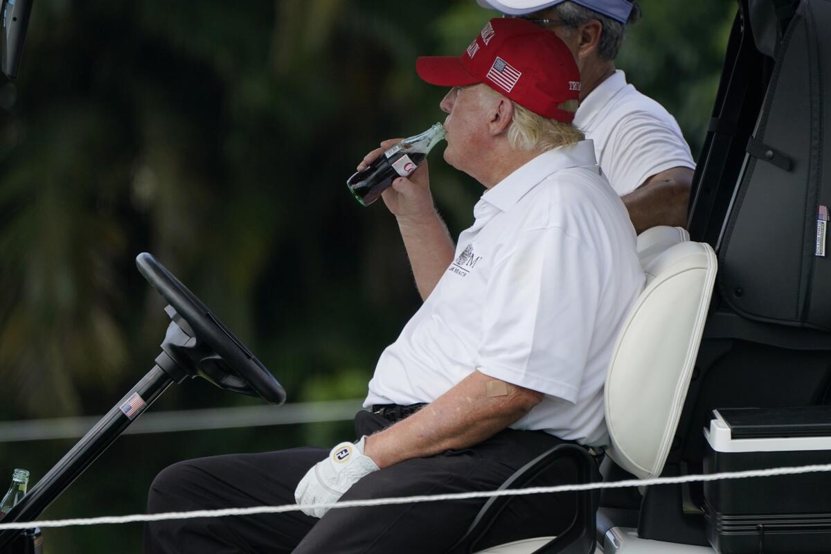 Former President Trump sitting in a golf cart drinking from a bottle of soda.