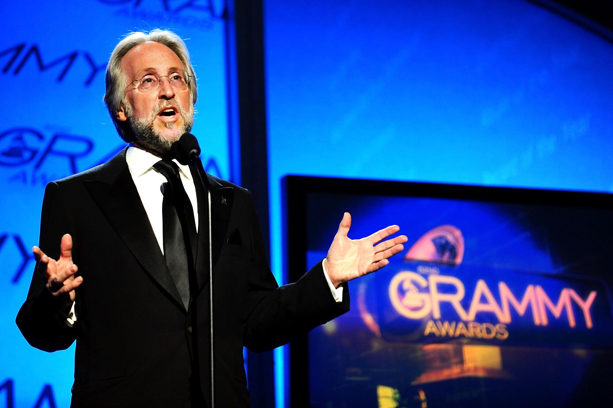 A bearded man in dark suit and tie stands at a mic and gestures. The words Grammy Awards are illuminated behind him.