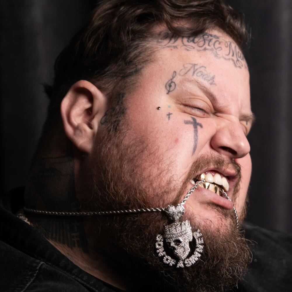 A country singer with face tattoos poses with a silver necklace in his mouth
