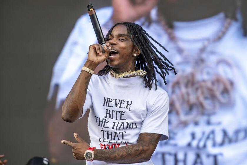 A young man with braids wearing chains, a white t-shirt and a watch rapping into a microphone on a stage