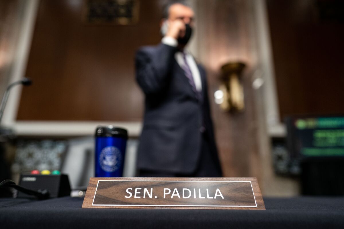 Sen. Alex Padilla speaks on the cellphone standing behind his seat in a committee hearing room. 