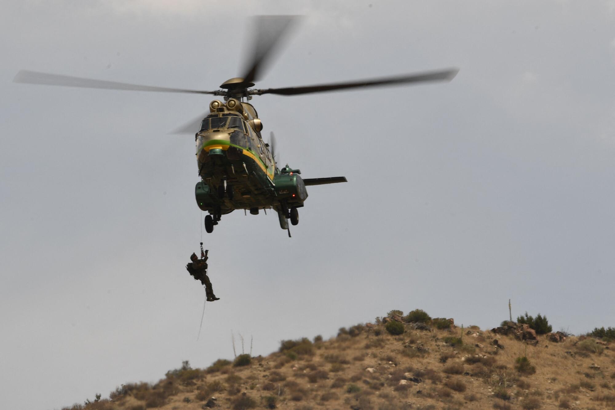 A man dangles from a cable attached to a helicopter hovering over a hillside.