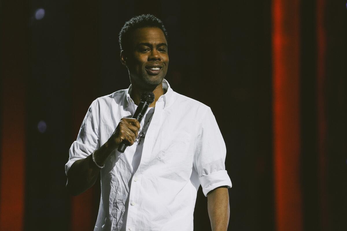 Chris Rock in a white button down shirt holding a microphone on a stage