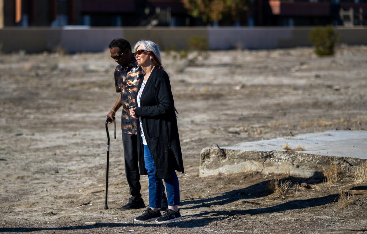 Two people, one with a cane, walking past a cement slab in the dirt of a vacant lot.