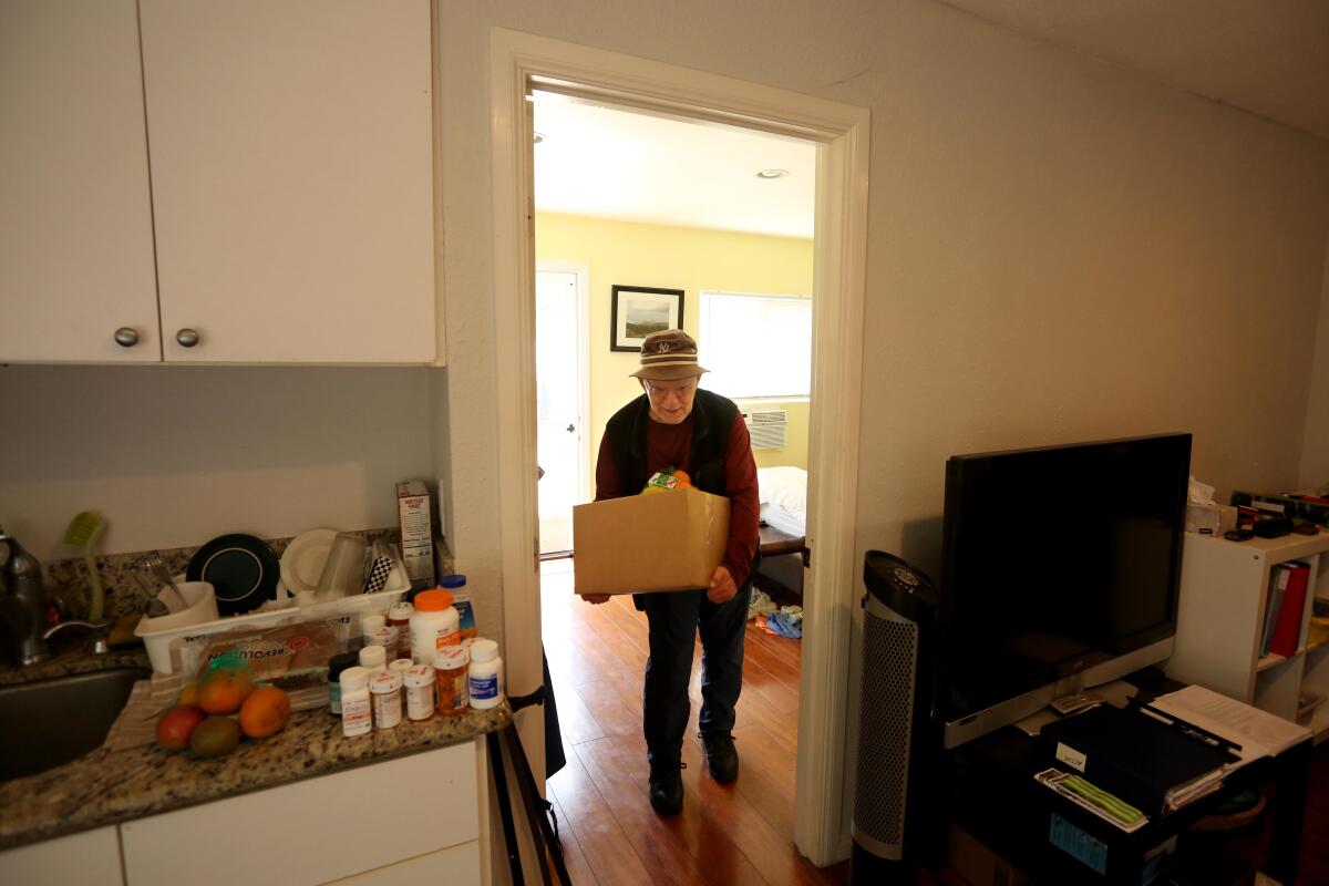 A man lugs a cardboard box brimming with food toward the kitchen of a small dwelling unit