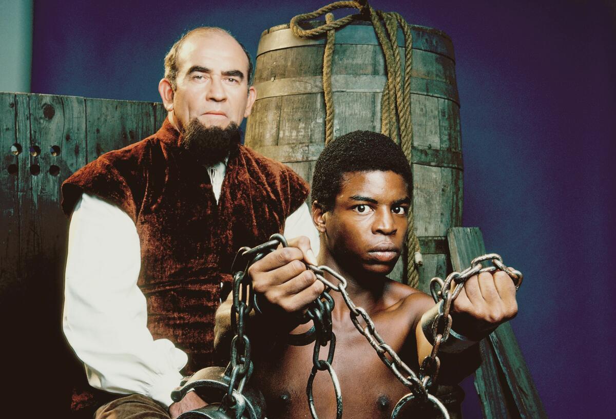 A white man in period garb stands with a black man in chains