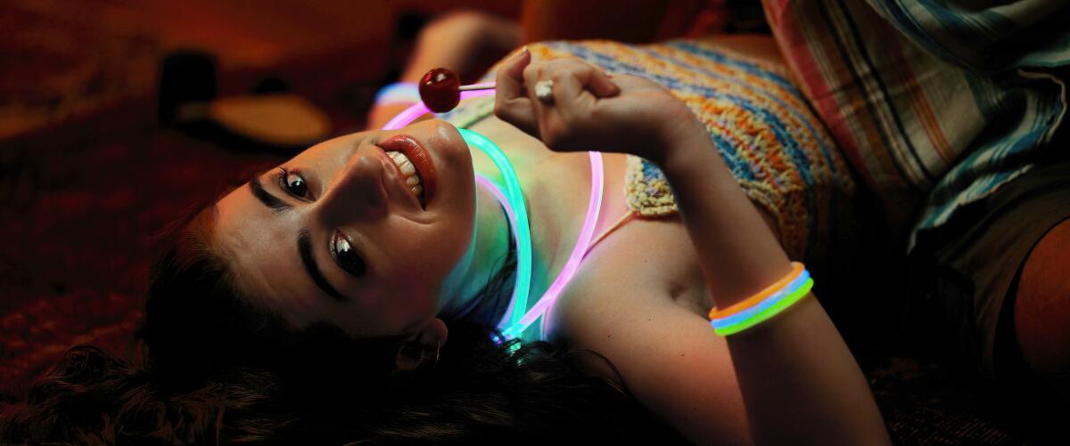 A young woman wearing glowing necklaces and holding a sucker lies on the floor.