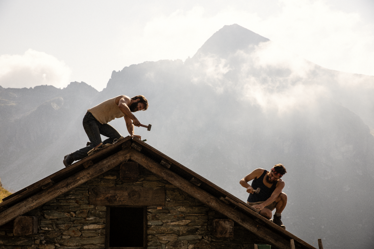 two men work on the roof of a house, with mountains in the background 