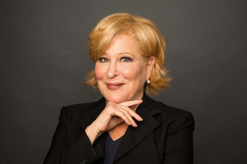 Bette Midler posing with one hand under her chin, clad in a black suit in front of a dark studio background