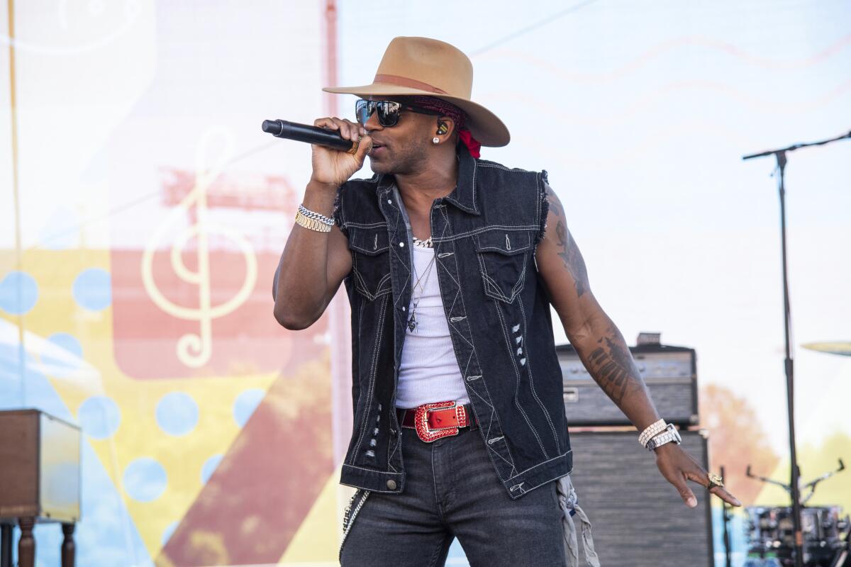Jimmie Allen performs onstage in a brown wide-brimmed hat and a jean vest