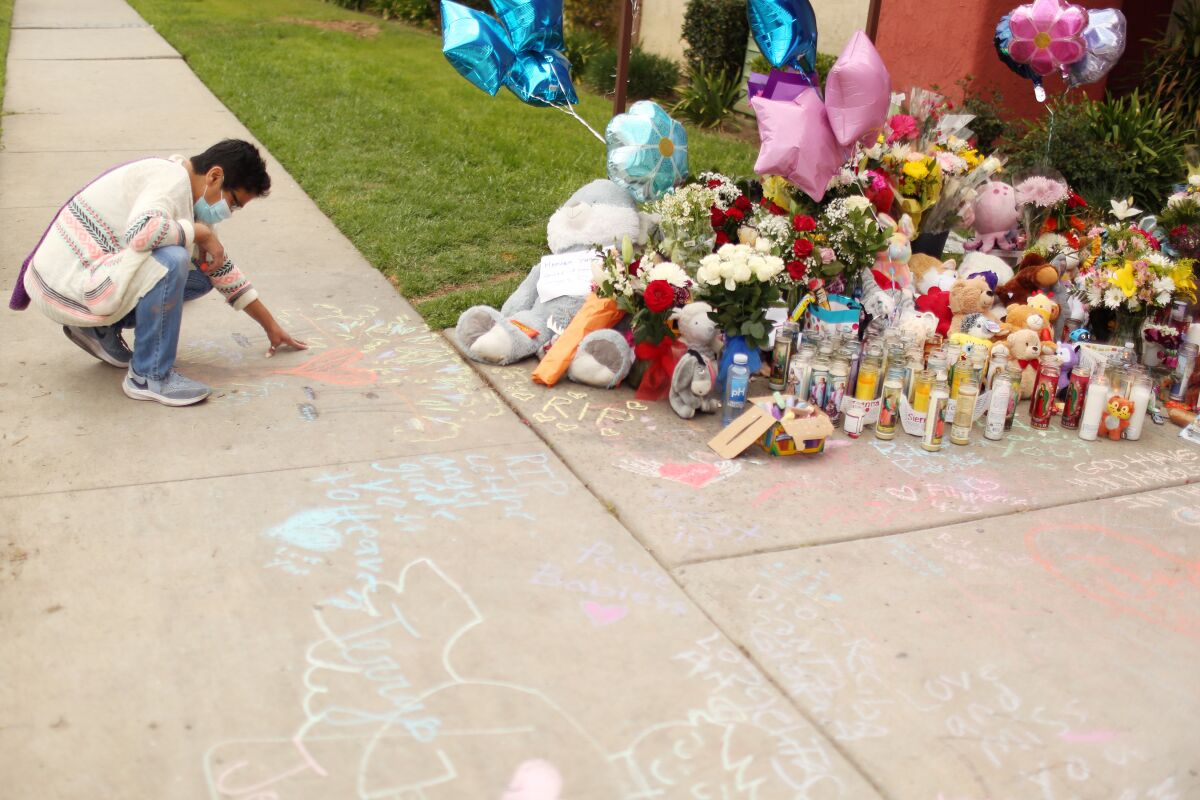 A woman kneels down on a sidewalk next to a memorial with balloons, flowers, candles and toys.
