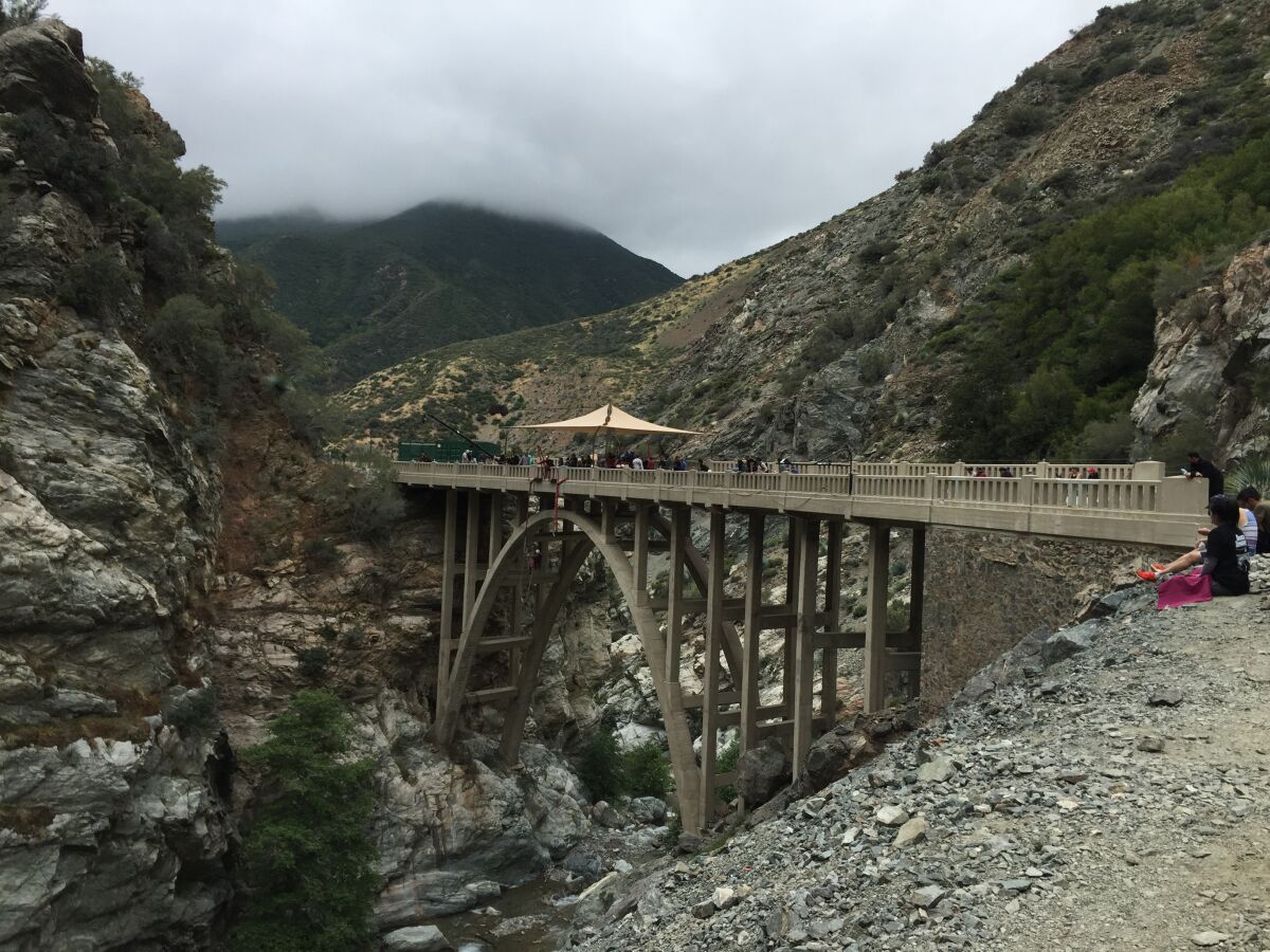 A large concrete bridge spans a mountainous area as people stand on the bridge.