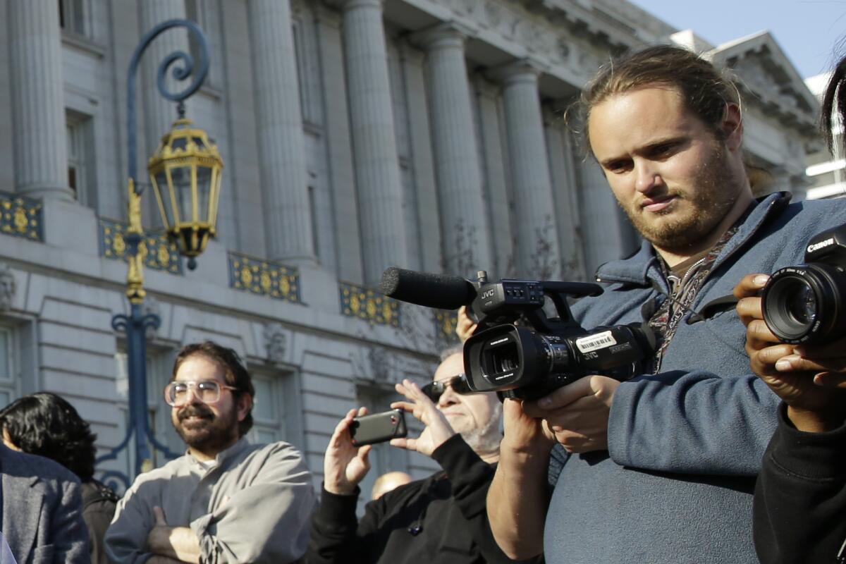 A man holds video camera outdoors among other people.