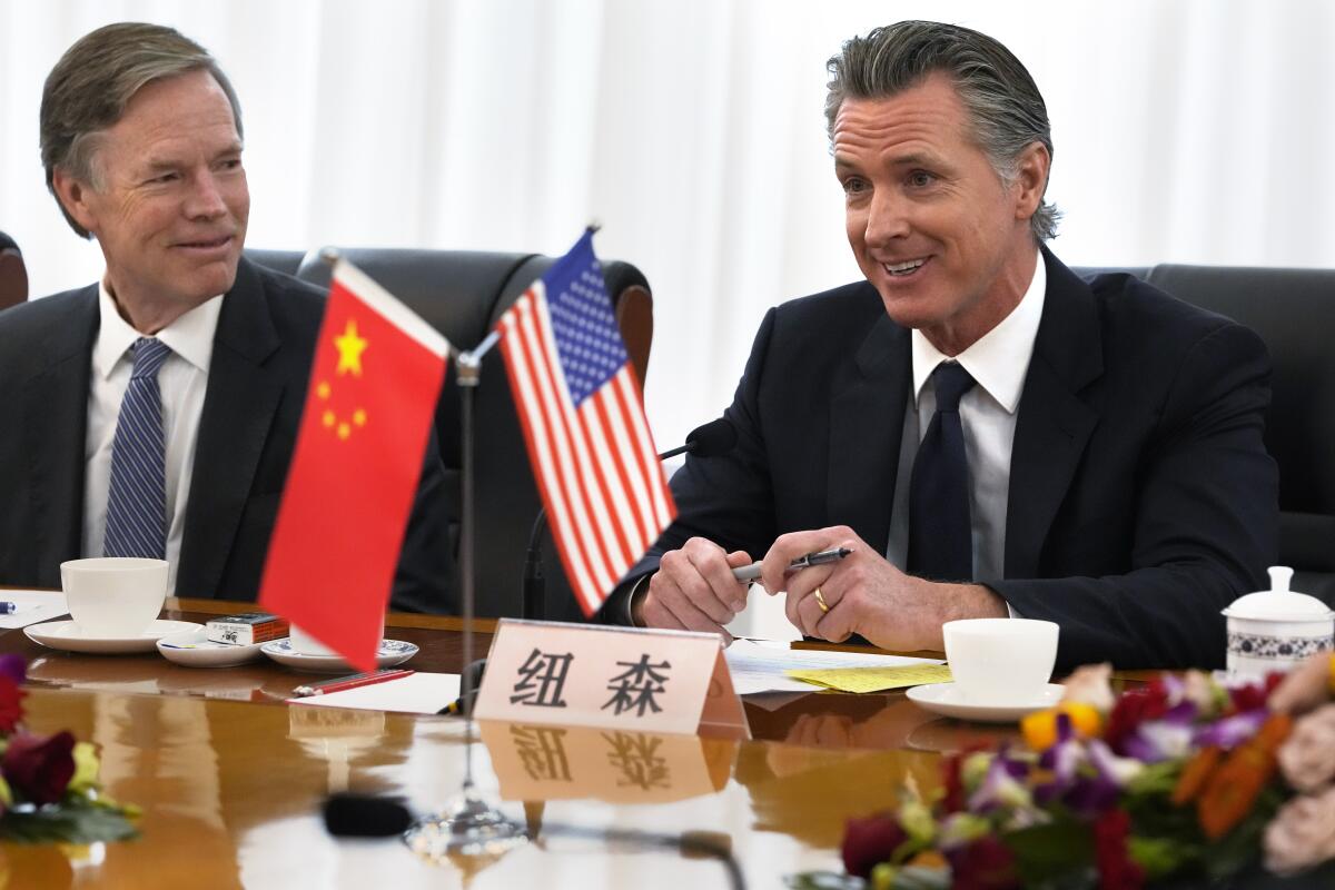 California Gov. Gavin Newsom smiles and sits at a table with small U.S. and Chinese flags and flowers next to another man.