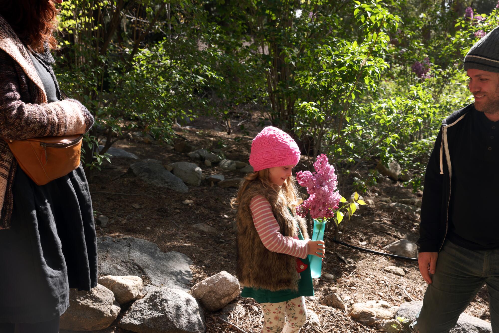 A young girl holds a bouquet of pink lilacs as two adults look on.