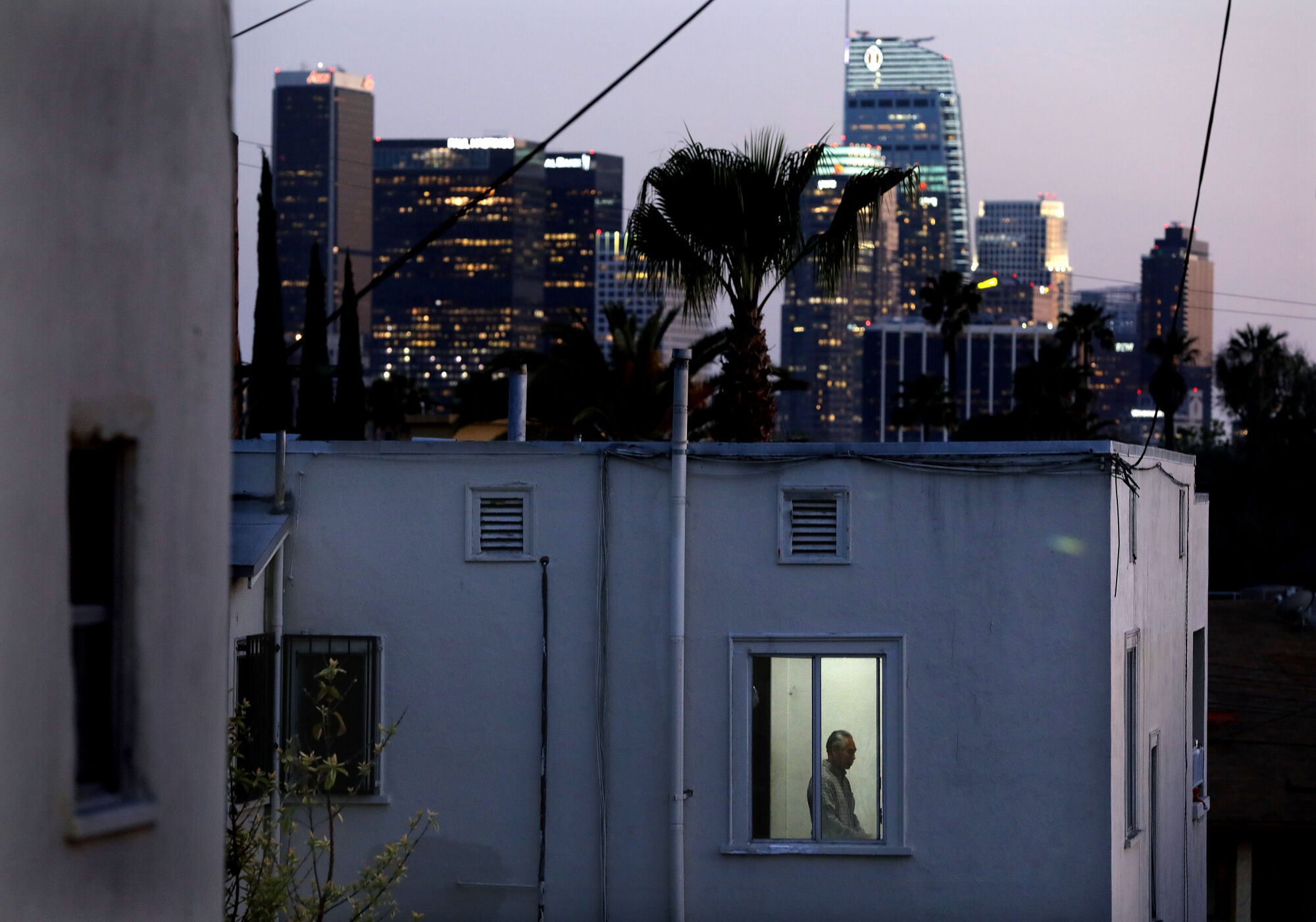 A man is framed in the window of an apartment, with a downtown L.A. in the distance. 