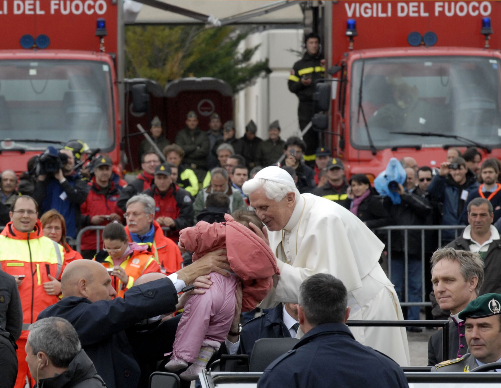 Papa Bento XVI  beija um bebê que está sendo segurado na frente dele ao ar livre enquanto várias pessoas estão olhando.