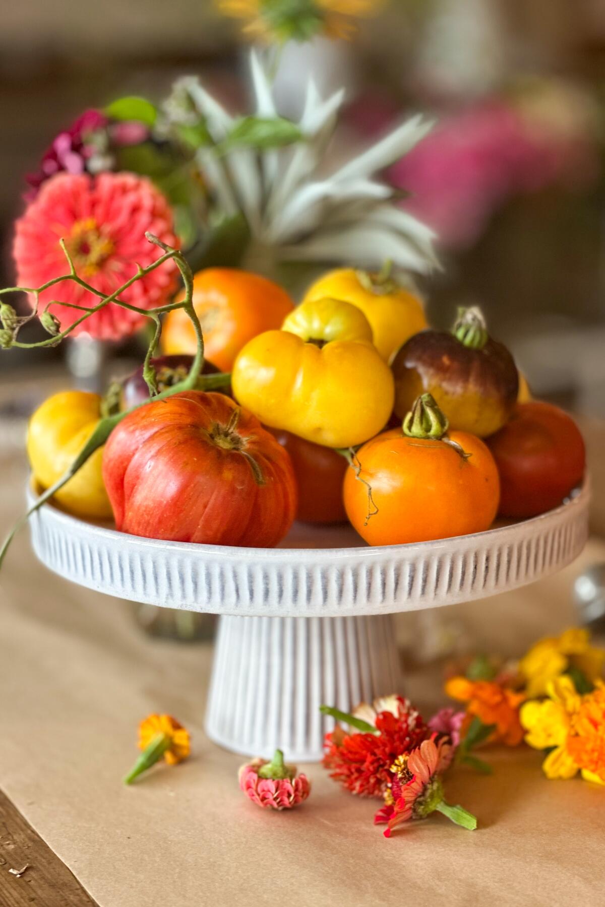 Red, yellow, orange and purple tomatoes piled on a white pedestal, studded with colorful zinnias.