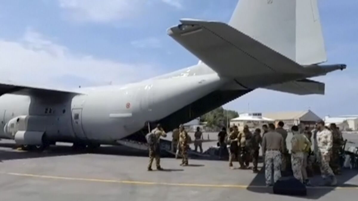 People in military fatigues stand under the tail of a military transport plane