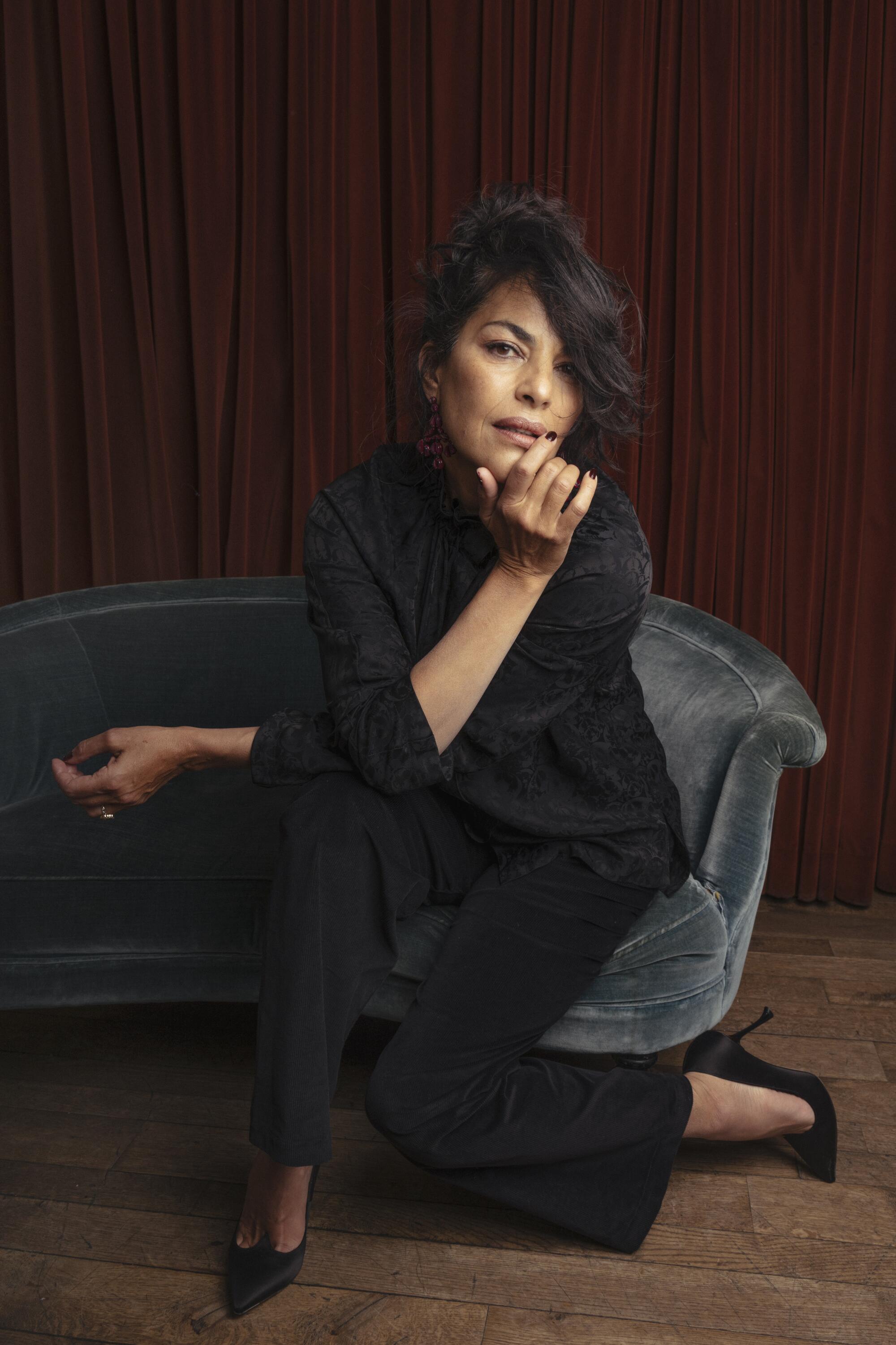 A woman in a black blouse, pants and heels poses on a small blue sofa