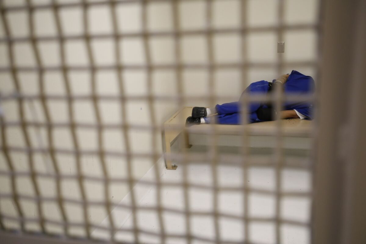 A detainee in blue clothing lies sideways on a jail bed