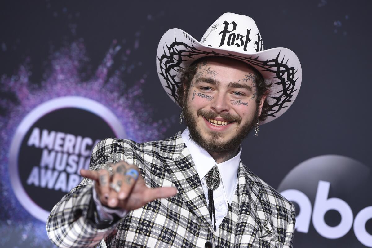 Post Malone is wearing a plaid suit with a bolo tie and white cowboy hat, smiling while gesturing.