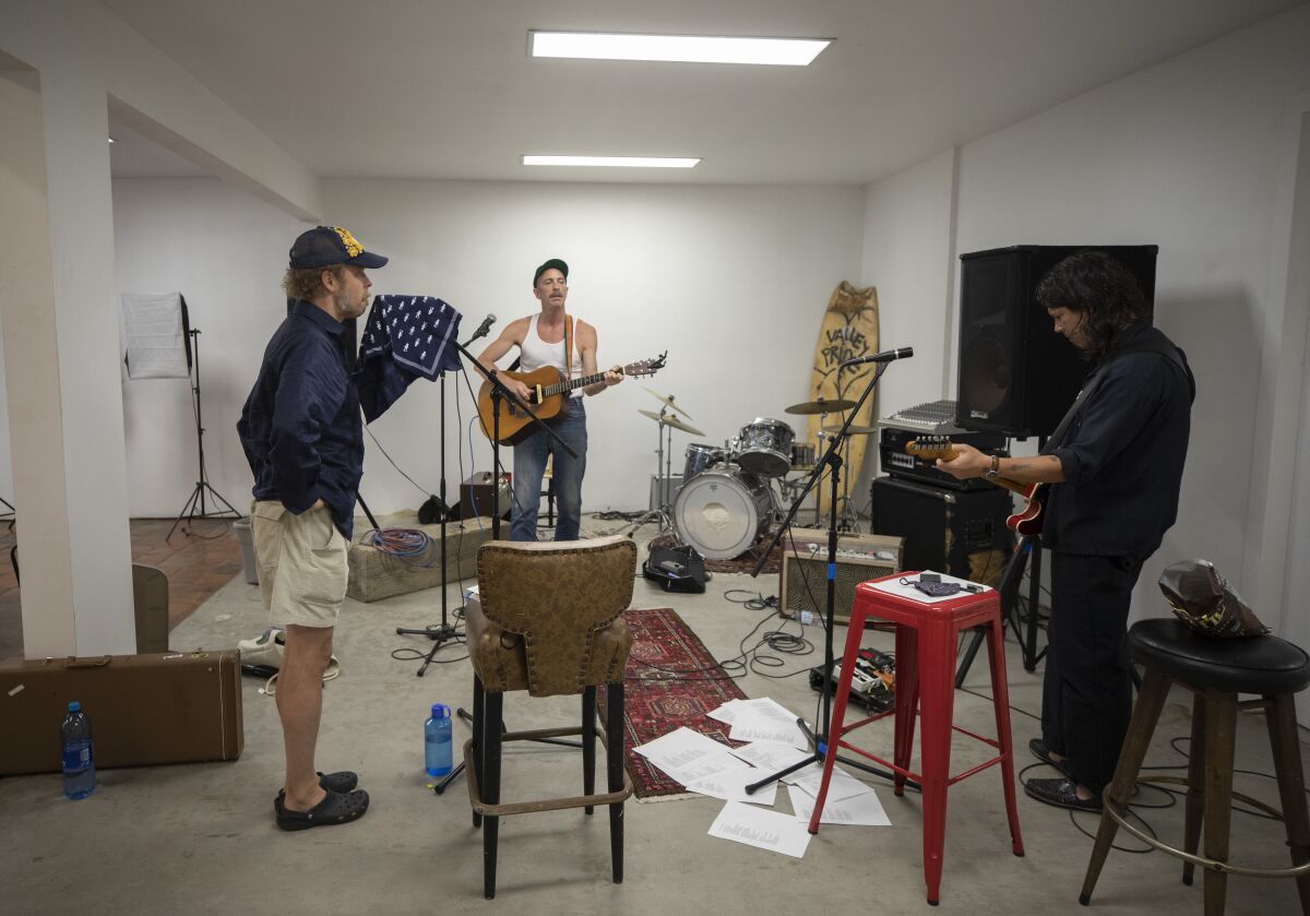 Three men stand facing each other in a room with papers scattered on the floor and a drumset and surfboard in the corner