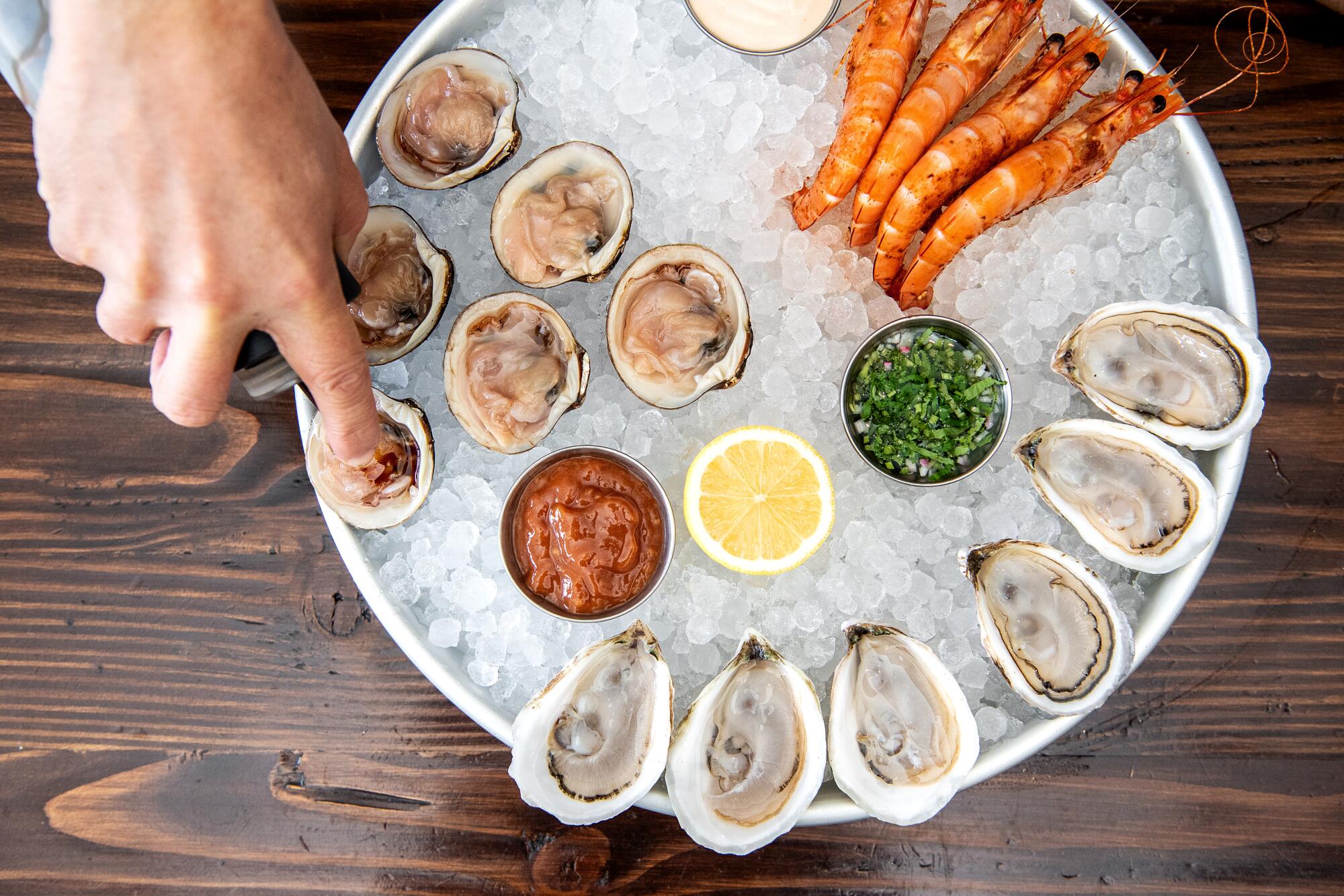 A chef's hand hovers over a tray of oysters, clams and prawns on ice.