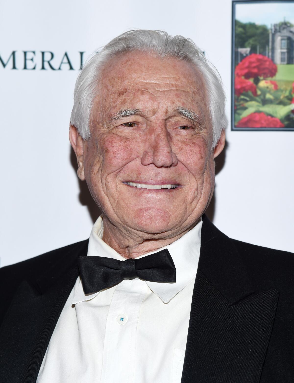 George Lazenby smiles and poses as he looks off camera while wearing a black and white tuxedo