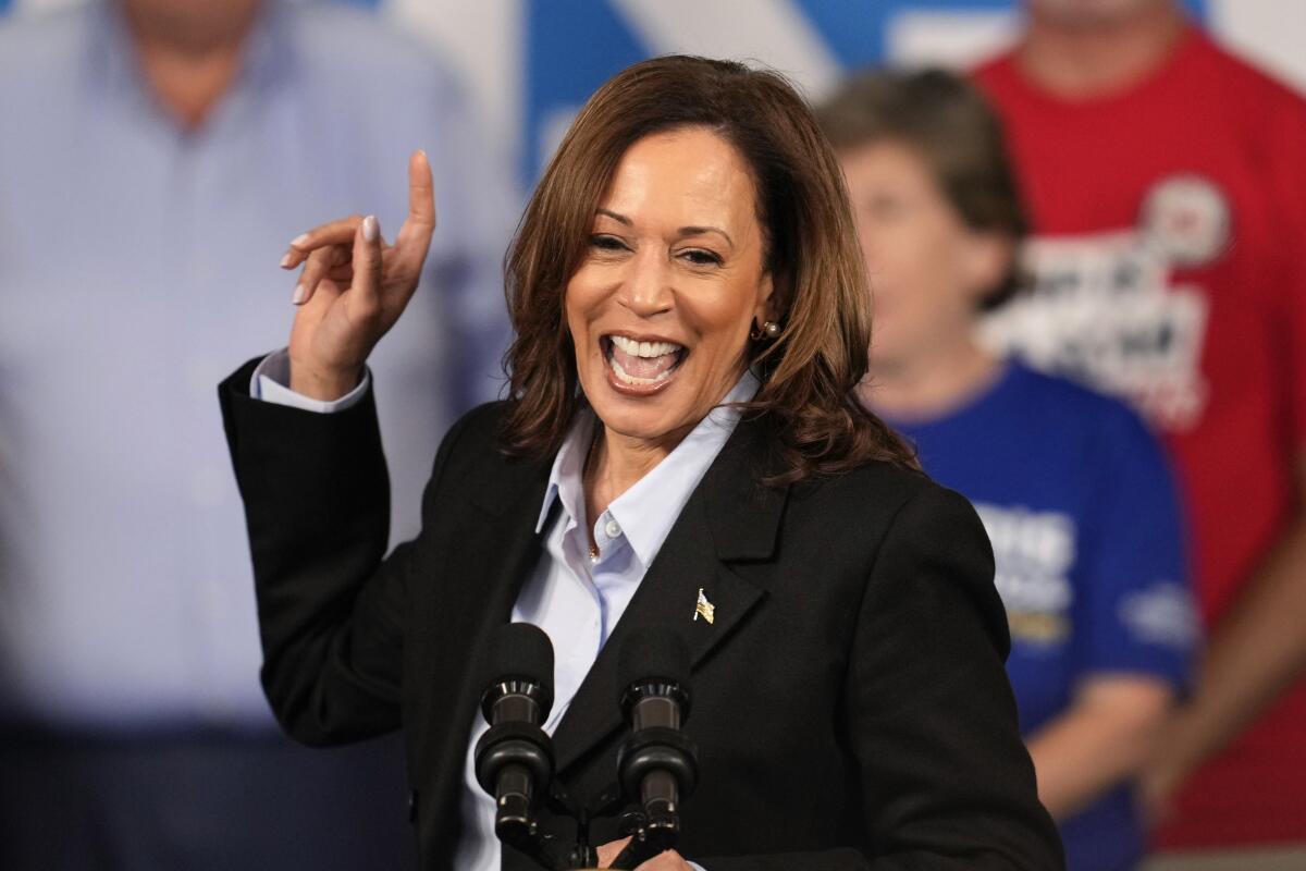 Kamala Harris stands smiling before a mic with one hand raised, finger pointed upward 