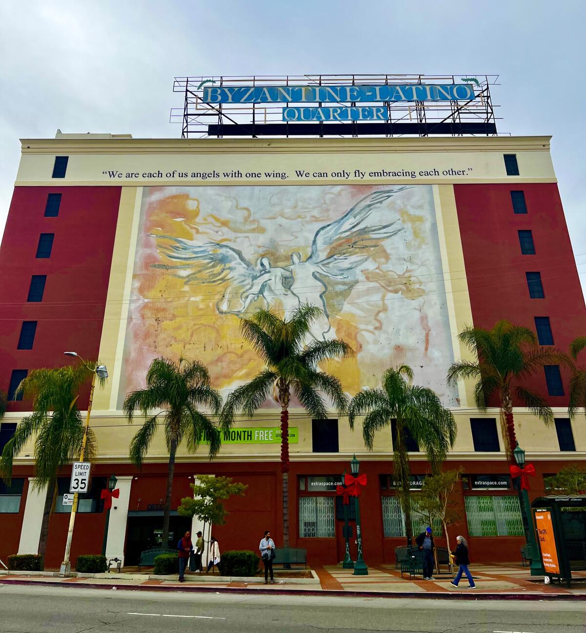 A large mural on a building depicts two people with one wing each, standing together to fly.