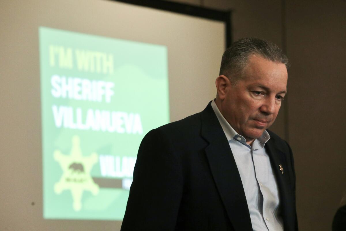 A man pauses while speaking before a sign that says, "I'm with Sheriff Villanueva"