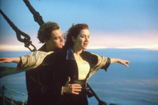 Leonardo DiCaprio places a his hands on the waist of Kate Winslet, whose arms are outstretched at the front of a ship.