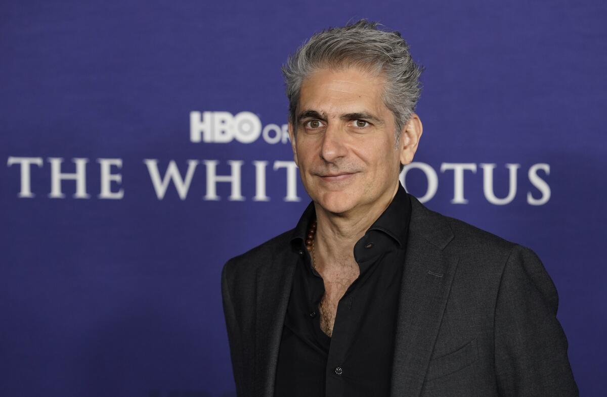 Michael Imperioli posing in a gray suit and black shirt against a purple background.