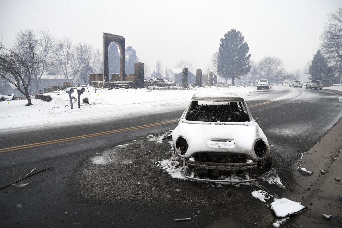A burned-out car, covered in a light layer of snow, sits in the middle of a road.