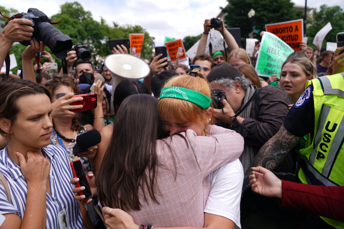 Women embrace within a crowd of people holding signs; media take photos.