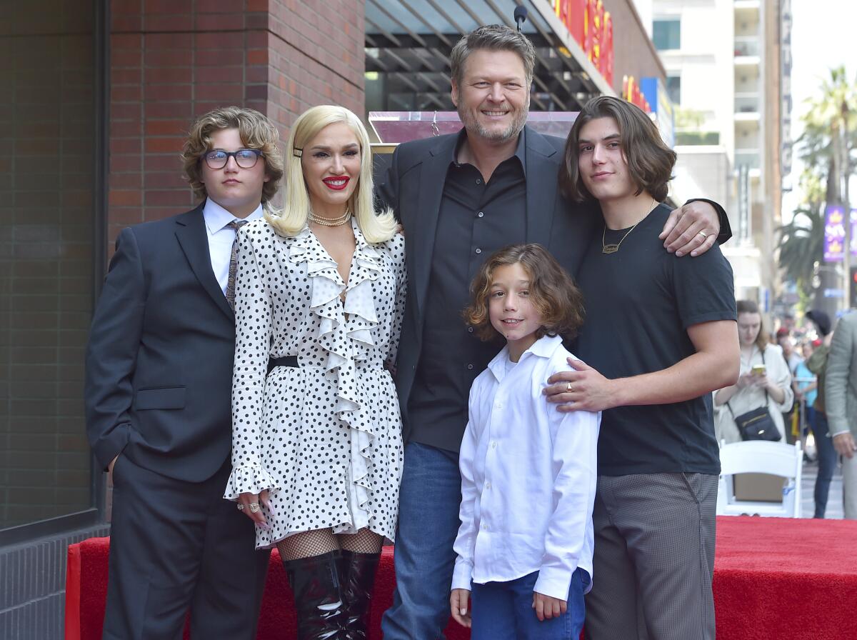 Gwen Stefani in a patterned white dress and Blake Shelton in jeans and a blazer with three young men outside a theater