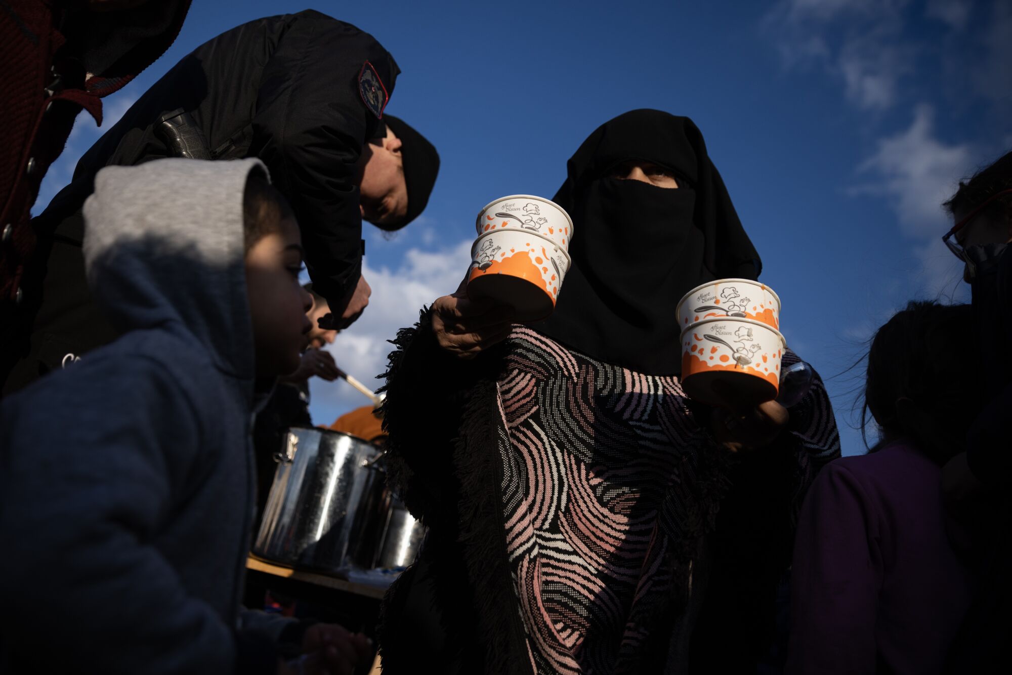 A woman balances several bowls as she stands in a food line near children.
