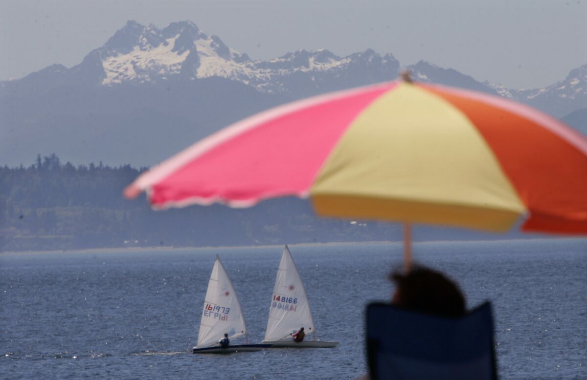 A pair of small sailboats float past beachgoers at Seattle's Golden Gardens Park. The Olympic Mountains are in the background.