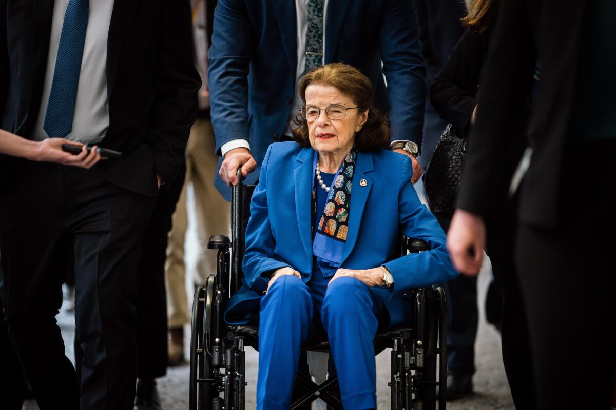Sen. Dianne Feinstein, wearing a blue pantsuit, sits in a wheelchair as others walk alongside her.