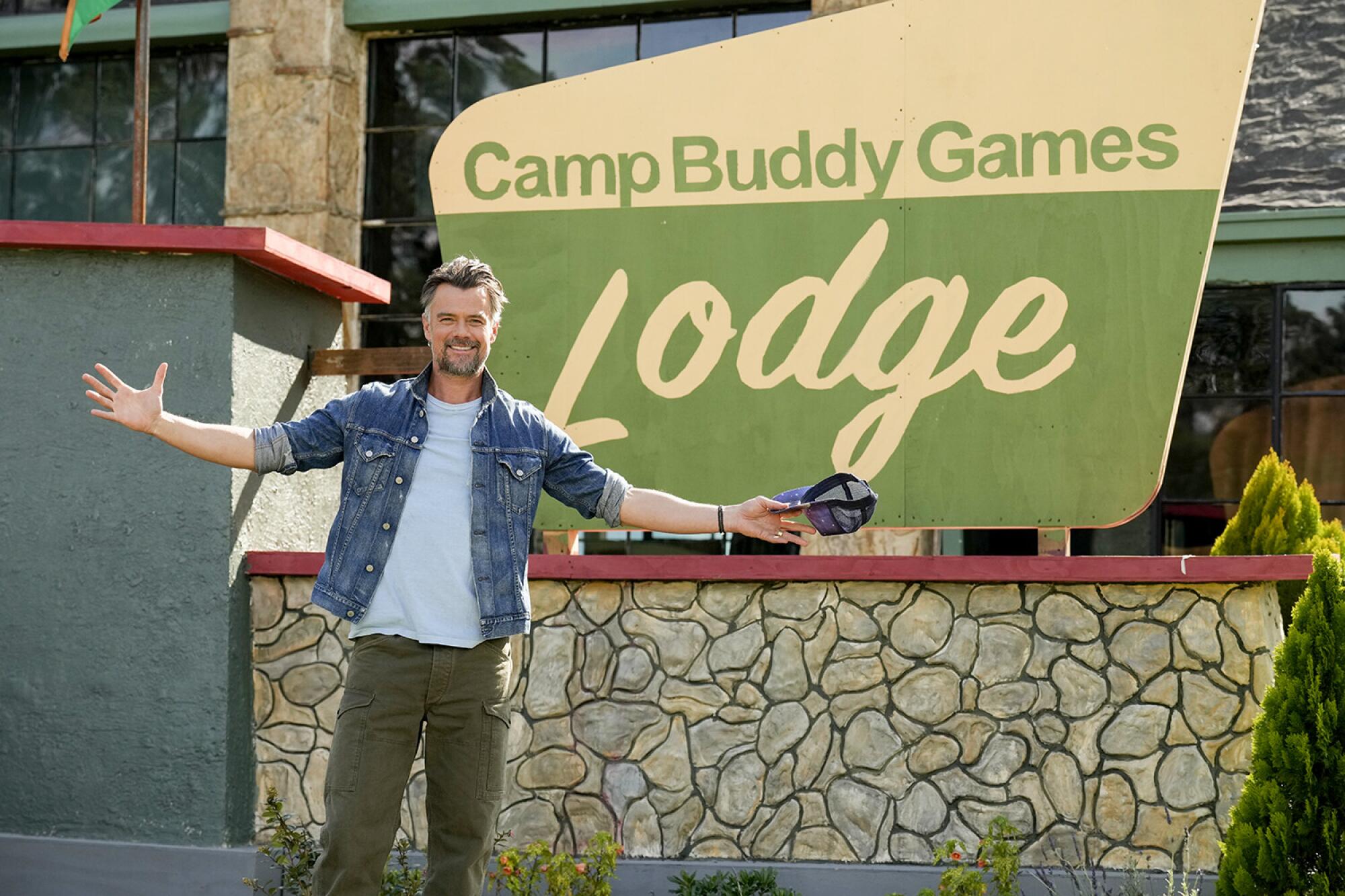Josh Duhamel stands with his arms open behind a sign that reads "Camp Buddy Games Lodge."