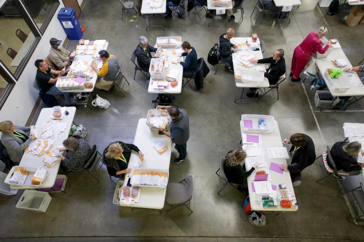 Overhead view of workers at long tables processing vote-by-mail ballots.