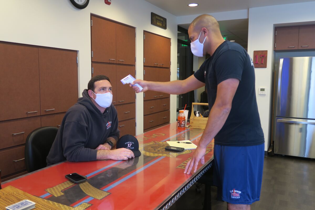 Capt. Dallas Higgins (standing) takes a temperature reading from engineer Dylan Surprise on Monday at station 17 in City Heights, the same day the San Diego Fire-Rescue Department implemented a cloth face covering policy for firefighters while in the station and out in public. Health screenings, including temperature readings, are performed twice daily and recorded.