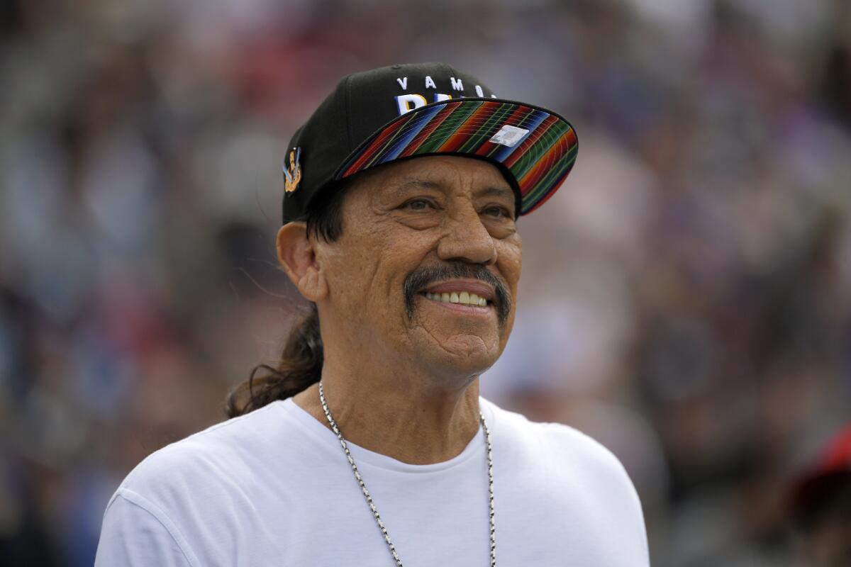 Danny Trejo smiling in a rainbow hat and white shirt.