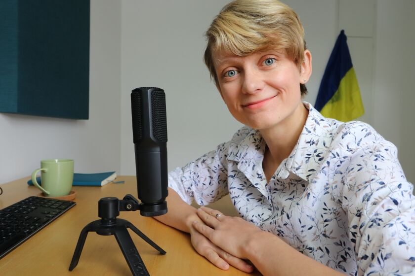 A woman with short blond hair sits smiling next to a microphone, with a Ukrainian flag in the background.