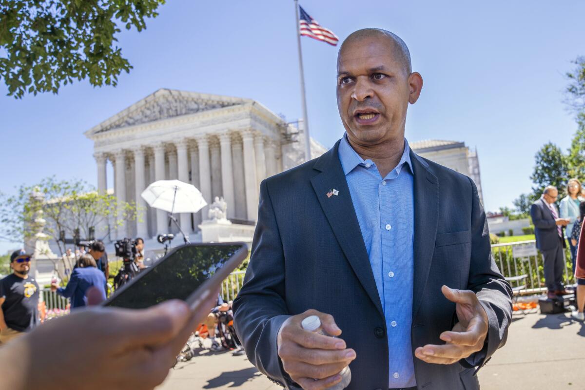 A man in a suit speaks to someone holding out a phone, with the U.S. Supreme Court building in the background.