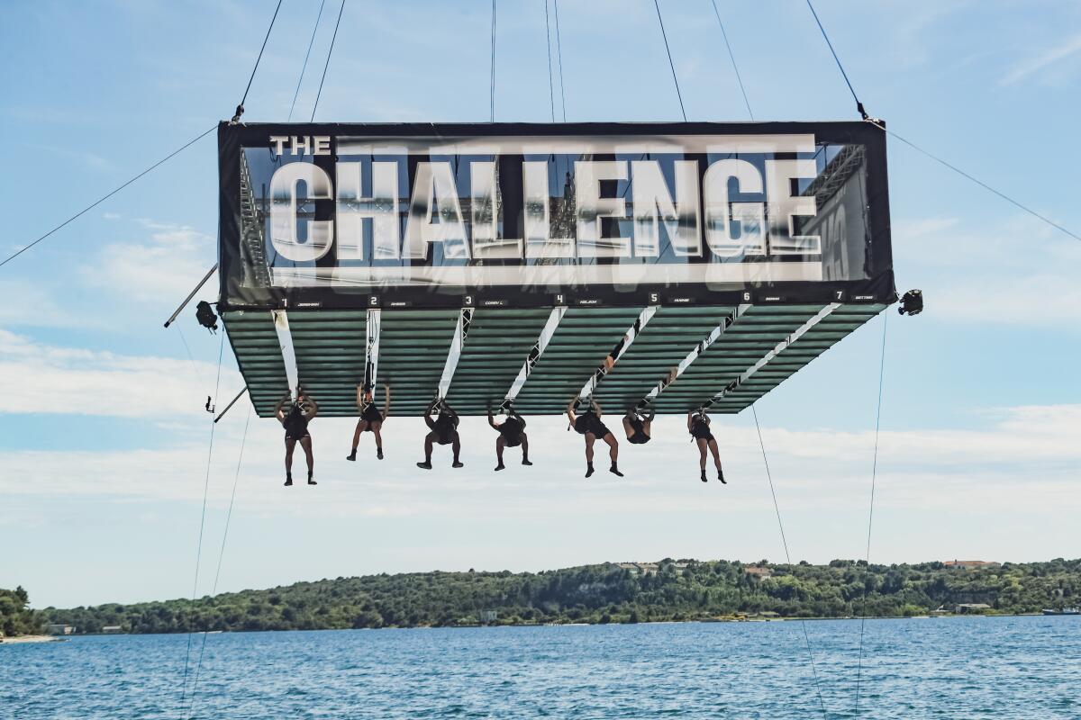 Competitors hang from a platform suspended over water labeled with the words "THE CHALLENGE"