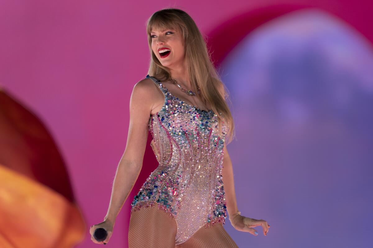 Taylor Swift smiles, poses and holds a microphone while wearing a sparkly bodysuit on a stage.