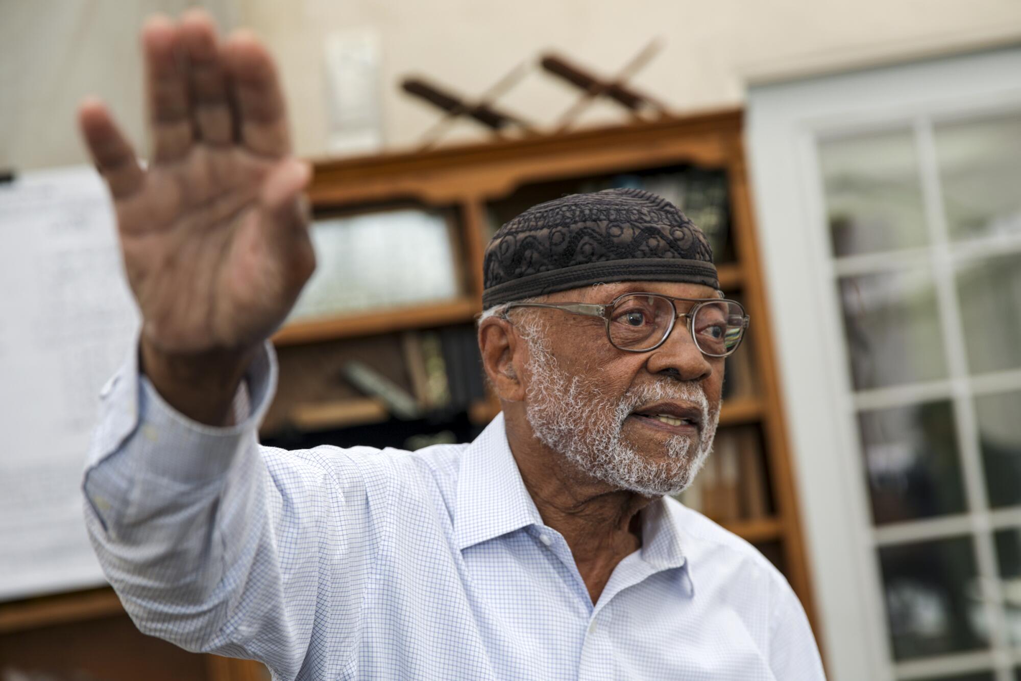 An older man wearing a cap and glasses with his hand raised