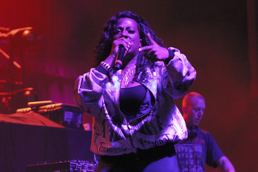 A woman with long black hair wearing a jacket and rapping into a microphone under purple lighting